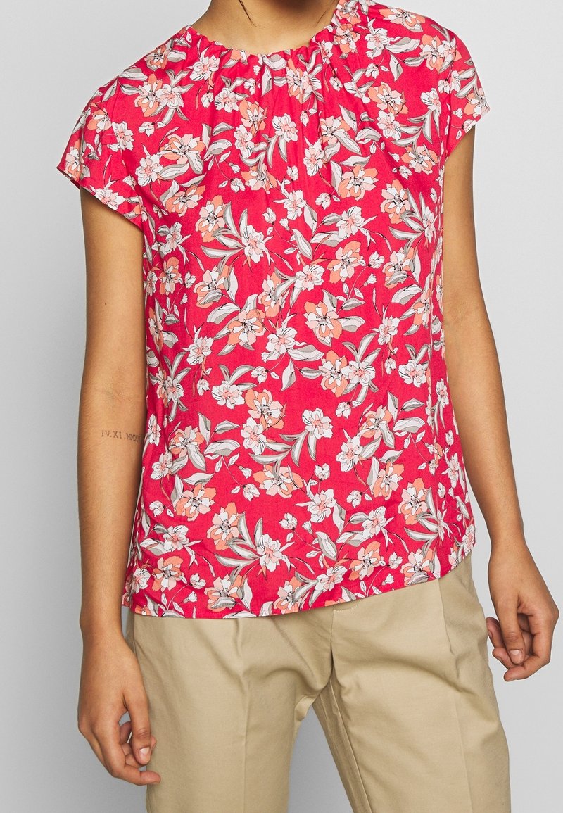 Bloemenblouse voor vrouwen in roze stof met korte mouwen en een geplooide halslijn. Het ontwerp bevat witte en koraalachtige bloemen met groene bladeren.