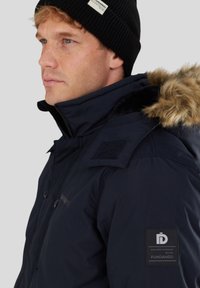 Veste isolante bleu marine avec une capuche en fourrure, patchs en Velcro sur les épaules et étiquette de logo sur le bras. Texture de tissu lisse et durable.