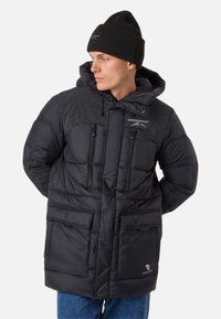 Black puffer jacket with a textured shoulder yoke, dual chest zippers, and front pockets. Paired with a black knit beanie.
