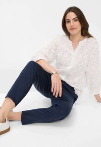 Witte geborduurde blouse met bloemenpatroon, lange mouwen en een kraaghals, gecombineerd met marina, taps toelopende broek en witte schoenen.
