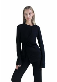 Melna ribota adīta blūze ar garām piedurknēm un sasietu priekšpusi. Materiāls ar teksturētu virsmu. Nēsāta kopā ar atbilstošām melnām biksēm.