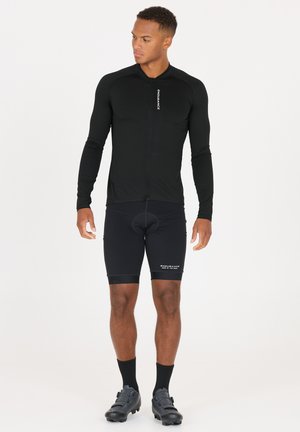 Atlet iført sort cykeltrøje med lange ærmer og polstrede shorts, sorte sokker og cykelsko, stående og kiggende til siden.