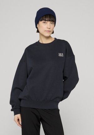 Osoba nosící tmavě modrou čepici a tmavě oversize mikinu s nápisem "Salt & Olives", ve spojení s černými kalhotami, na jednobarevném šedém pozadí.