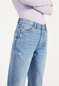 Persona vistiendo jeans de tiro alto desgastados de color azul claro y una camiseta de manga larga acanalada lila con los brazos cruzados frente a un fondo neutro.