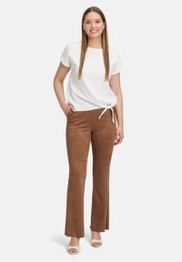 Biała bluzka z krótkim rękawem z bocznym węzłem, połączona z brązowymi zamszowymi spodniami dzwonami o gładkiej fakturze i minimalistycznym wzornictwie.