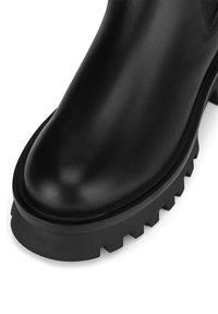 Czarny skórzany botek za kostkę z zaokrąglonym noskiem, grubą gumową podeszwą o postrzępionym wzorze i gładką fakturą.