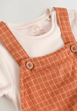 Bommullsbyxor i bränd orange med ett rutmönster, med träknappar och en krämfärgad skjorta under.