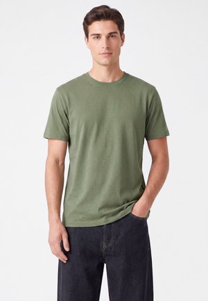Man draagt een effen olijfgroen T-shirt met korte mouwen en donkerblauwe jeans, staand met één hand in de zak tegen een witte achtergrond.