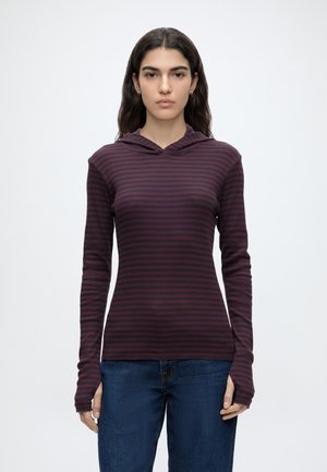 Jeune femme aux longs cheveux foncés, portant un haut moulant à capuche rayé bordeaux et noir à manches longues et un jean bleu, debout sur fond blanc.