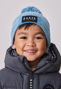 Modra pletena kapa z cofom na vrhu, z črno etiketo z napisom "BAKER BY TED BAKER." Kombinirana s sivo podloženo jakno.