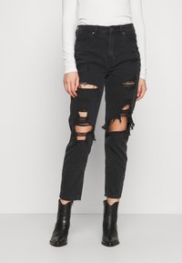 Jean noir déchiré avec de grandes déchirures et des bords effilochés, en denim. Associé à un haut à manches longues blanc et des bottines noires.