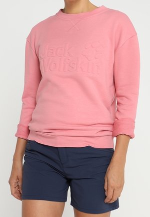 Osoba nosząca różową bluzę z podwiniętymi rękawami i wytłoczonym logo „Jack Wolfskin”, w połączeniu z granatowymi szortami.