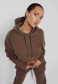 Brauner, verkürzter Hoodie mit Taschen auf der Vorderseite, Kordelzug und gerippten Bündchen; dazu passende braune Jogginghosen mit Logodetail; glatte Stofftextur.