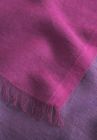 Écharpe violette avec un tissage fin et texturé. Les bords frangés ajoutent des détails, superposée à un tissu texturé plus foncé dans des tons de violet complémentaires.