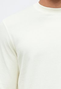 Hellcremefarbenes Sweatshirt mit glatter Textur, geripptem Rundhalsausschnitt und leicht tailliertem Schnitt, ohne sichtbare Muster oder Akzente.