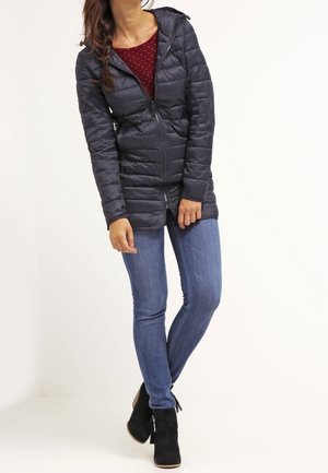 Kobieta w czarnej pikowanej kurtce z kapturem, czerwonej koszuli w białe grochy, niebieskich obcisłych dżinsach i czarnych botkach, stojąca z lekko ugiętymi nogami.