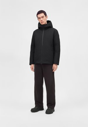 Homme portant une veste à capuche noire, un pantalon noir et des chaussures noires, se tenant devant un fond gris uni.