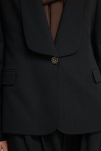 Zwarte blazer met een pieklapel, krijtstreep patroon en een metalen knoop. Gestructureerde stof en twee voorzakken zijn zichtbaar.