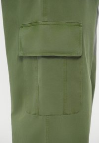 Πράσινα cargo παντελόνια με μεγάλη τσέπη κλειστού τύπου, λείες υφές και διπλοραμμένα ραφάκια, τονίζοντας την πρακτικότητα στο σχέδιο.