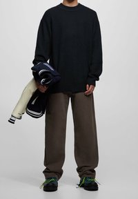 Pull en maille côtelée vert foncé, pantalon ample marron, portant une veste marine et un autre article marine, avec des chaussures noires à lacets verts.