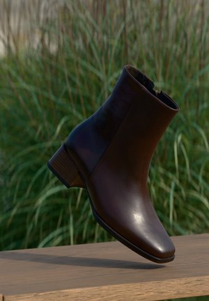 Donkerbruine leren enkellaarsjes met een lage blokhak die boven een houten oppervlak zweven, met op de achtergrond wazig hoge groene gras.