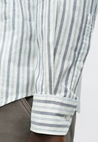 Napillinen paita, jossa pystysuorat sinivalkoiset raidat, puuvillamateriaalia. Paidassa taitettava kalvosin ja kankaassa on kevyt kiilto.