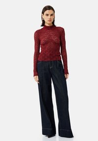 Top long-sleeve rosso in pizzo con collo alto, abbinato a jeans in denim scuro a vita alta e gamba larga, con dettagli di cucitura in vista.