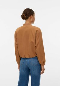 Femme avec un chignon de cheveux bruns portant un haut à manches longues marron et un jean bleu, de dos contre un fond blanc uni.