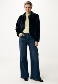 Svart fuskpälsjacka med tryckknäppning, bärs över en ljusgrön polotröja. Matchas med vida blå denimjeans och svarta skor.