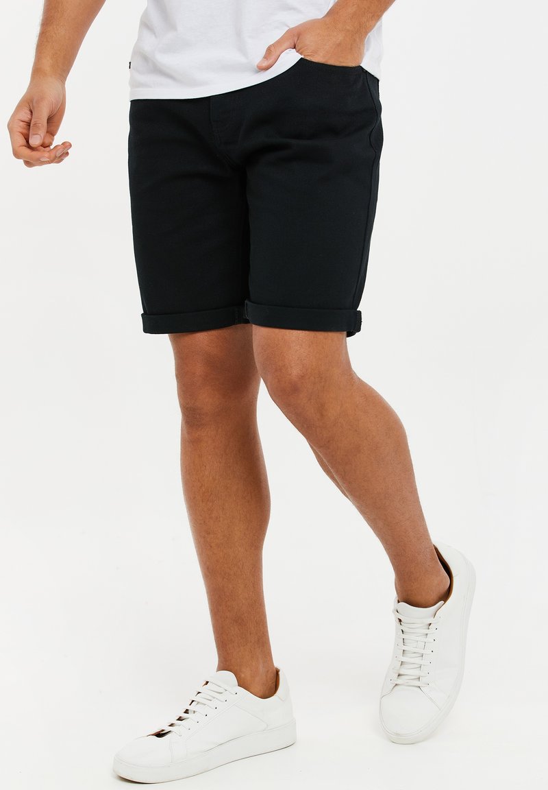 Shorts en coton noir avec des revers roulés, poches latérales et une coupe décontractée, associés à des baskets blanches, avec un système de laçage et des semelles texturées.