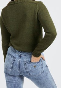 Personne portant un pull en tricot olive et un jean bleu clair délavé, avec une main dans la poche arrière, sur un fond uni.