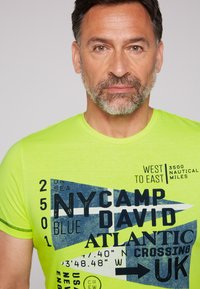 Camiseta amarilla neón con estampado gráfico que presenta texto en azul y negro, formas geométricas y temas náuticos. Diseño de manga corta y cuello redondo.