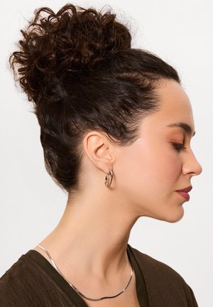 Srebrny kolczyk w formie obrączki oraz zakrzywiony metalowy naszyjnik noszony przez osobę z kręconymi ciemnymi włosami. Ubranie wykonane jest z teksturowanej brązowej tkaniny.