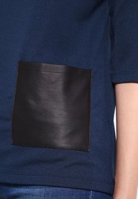 Námořnická modrá krátká rukávová blůza s velkou černou koženou náprsní kapsou vpředu. Měkká textura, volný střih, jednoduché švy.