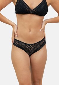 Ensemble de lingerie en dentelle noire comprenant un soutien-gorge triangulaire avec des bords festonnés et un délicat nœud, ainsi que des culottes taille basse assorties avec des motifs complexes.
