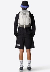 Zaino in tessuto grigio chiaro con schiena imbottita, tasche laterali in rete e spallacci regolabili. Indossato con felpa nera e pantaloni corti, calzature grigie.