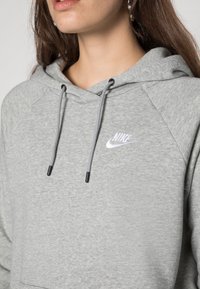 Šedá mikina s texturovanou tkaninou, nastavitelnou šňůrkou a vyšitým bílým logem Nike na hrudi. Má raglánové rukávy.