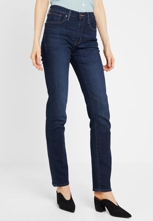 Frau trägt dunkelblaue, schmal geschnittene Jeans und schwarze Blockabsatzschuhe mit spitzer Spitze vor einem schlichten weißen Hintergrund.