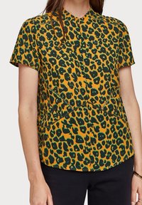 Blouse à manches courtes en tissu jaune moutarde, ornée d'un imprimé léopard vert, avec un col mandarin et des détails de couture inclinés.