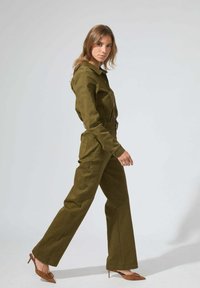 Combinaison verte olive à manches longues et fermeture à boutons, dotée d'une taille ceinturée et de deux poches latérales, accompagnée de chaussures à talons marron.