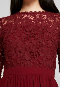 Burgundské šaty s krajkovým korzetem, který zdobí květinové vzory, dlouhé rukávy a nařasenou sukni s hladkým pasem.