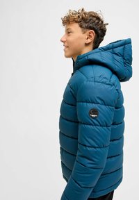 Tyrkysová puffer bunda se zipem a kapucí. Má prošívanou texturu a kulatou černou nášivku na levém rukávu. Hladký, izolační materiál.