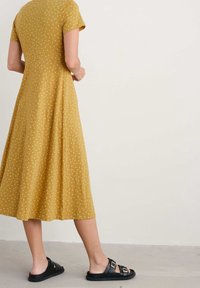 Sukienka z krótkim rękawem w kolorze musztardowym w białe kropki, w fasonie A, z miękkiego materiału, zestawiona z czarnymi sandałami z klamrami.