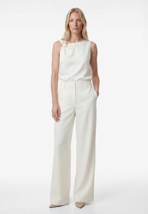 Kobieta o blond włosach, ubrana w białą, bezrękawnikową bluzkę z złotym detalem w kształcie pierścienia i szerokie białe spodnie, stojąca z jedną ręką w kieszeni.