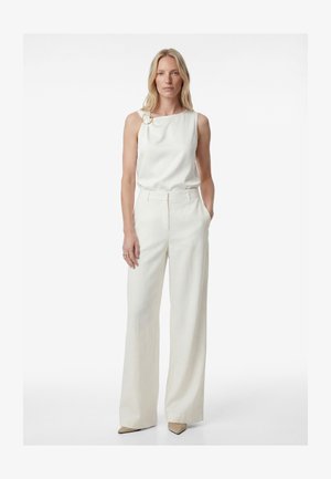 Kobieta o blond włosach, ubrana w białą, bezrękawnikową bluzkę z złotym detalem w kształcie pierścienia i szerokie białe spodnie, stojąca z jedną ręką w kieszeni.