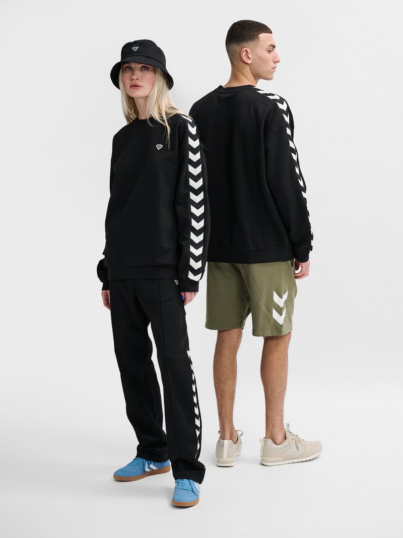 Deux modèles portent des sweats noirs avec des bandes blanches en chevron sur les manches ; l’un en pantalon noir et chaussures bleues, l’autre en short olive et baskets beiges.