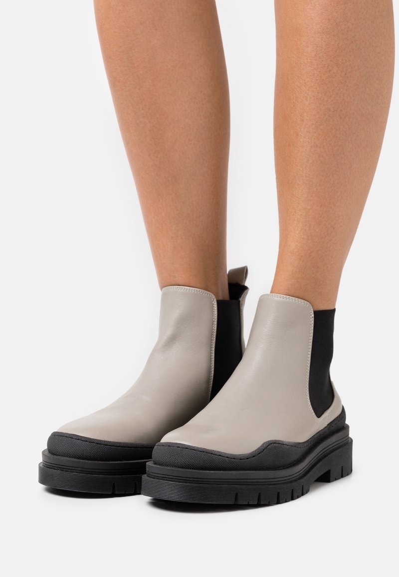 Bottes en cuir grises montantes à la cheville avec côtés élastiques noirs et semelles épaisses à crampons noirs, portées sur jambes nues.