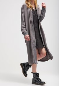 Lång grå kimono-liknande kappa i slät tyg med rak passform, öppen front och elastiska manschetter, kombinerad med en svart klänning och blanka svarta stövlar.