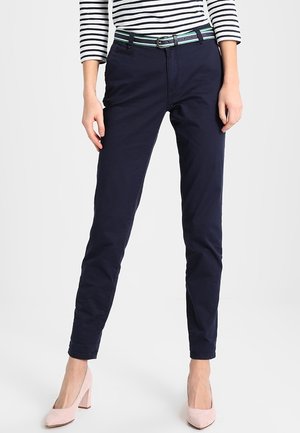 Frau trägt marineblaue Slim-Fit-Hosen, ein schwarz-weiß gestreiftes langärmliges Oberteil, grünen Gürtel und blassrosa High Heels, stehend.