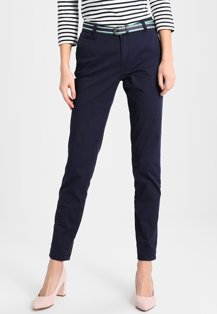 Femme portant un pantalon slim bleu marine, un haut à manches longues rayé noir et blanc, une ceinture verte et des chaussures à talons hauts rose pâle, debout.